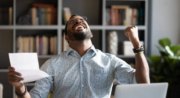 Overjoyed young mixed race man in glasses holding paper in hands, celebrating unbelievable triumph, sitting at workplace. Excited happy millennial african american businessman received loan approval.